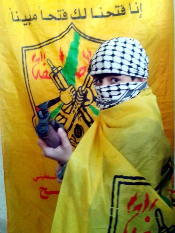 A Gaza child draped in a Fatah flag poses with a weapon.