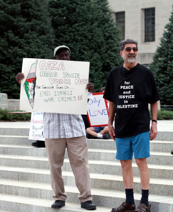 An anti-Israel protester holds a sign falsely accusing Israel of genocide and war crimes. The map on the sign reveals the ambition to wipe out Israel and for a Palestinian state to replace it. The sign behind him on the left tells passersby to boycott Israel.