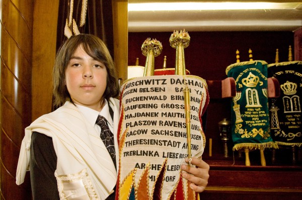 A Jewish teen holds a Torah scroll covered by an ornately decorated Torah mantle that commemorates those who perished in Nazi death camps during the Holocaust.