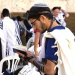 A Jewish man wearing a kippah, tefillin (phylacteries), and a tallit (prayer shawl) prays at the Western (Wailing) Wall using a siddur (prayer book).