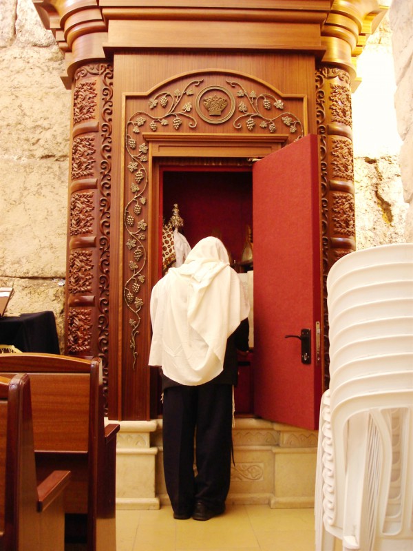 A Jewish man with his tallit (prayer shawl) draped over his head stands before an open Torah ark at the Western (Wailing) Wall.