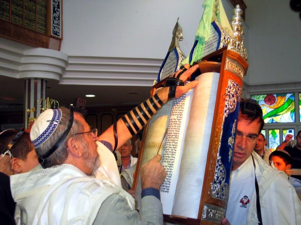 A Jewish man wearing tefillin (phylacteries) and a tallit (prayer shawl) reads from an open Torah scroll housed in an ornate Torah tik.